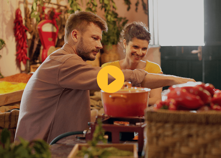 Male and female both cooking over a stove with a red/orange pot. Red peppers hanging in the background